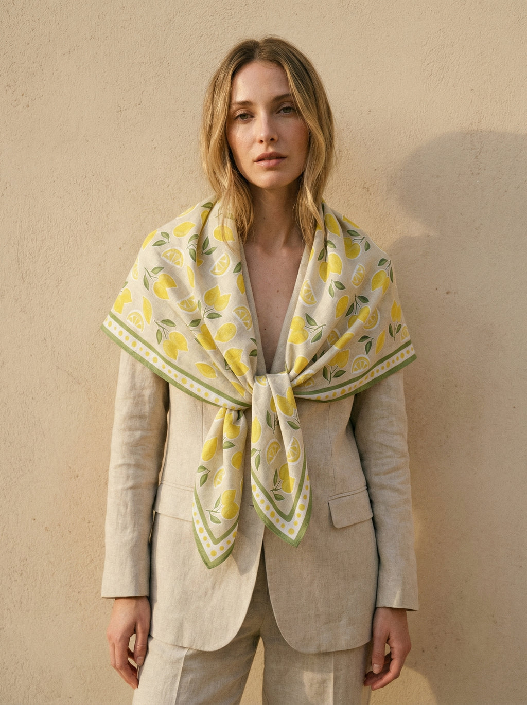 Model folding a Maison OYO 100% linen square scarf Limoncello design, yellow lemon pattern and green leaves on natural linen background, green border with yellow polka dots.